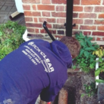 A drainage engineer investigating a residential drain beside a brick wall. Image sourced from the Quick Clear Drainage website.