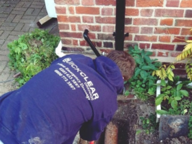 A drainage engineer investigating a residential drain beside a brick wall. Image sourced from the Quick Clear Drainage website.