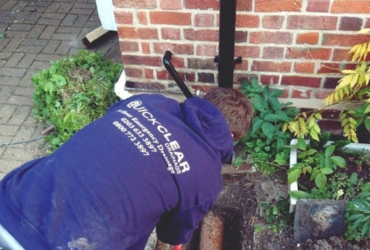 A drainage engineer investigating a residential drain beside a brick wall. Image sourced from the Quick Clear Drainage website.