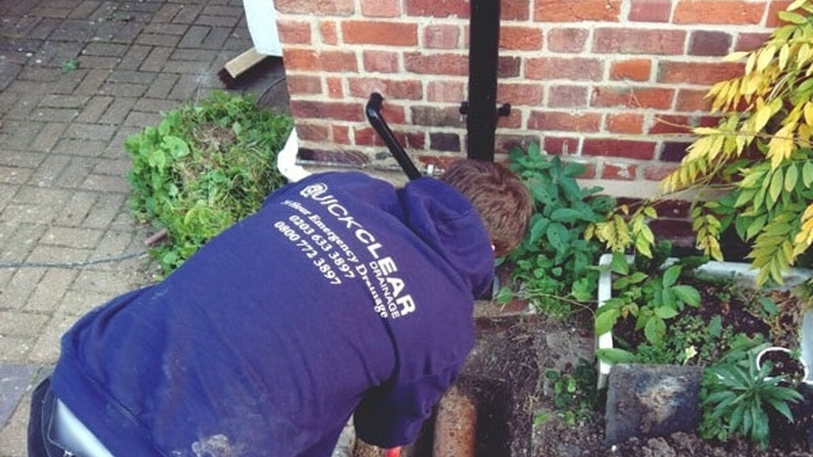 A drainage engineer investigating a residential drain beside a brick wall. Image sourced from the Quick Clear Drainage website.